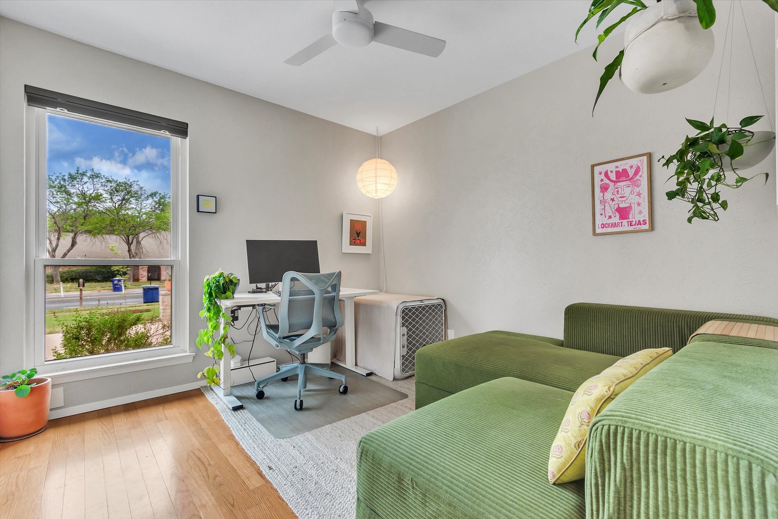 Bedroom 2 used as home office — green corduroy sofa, desk with monitor, hanging plants, hardwood floors, and large window with natural light