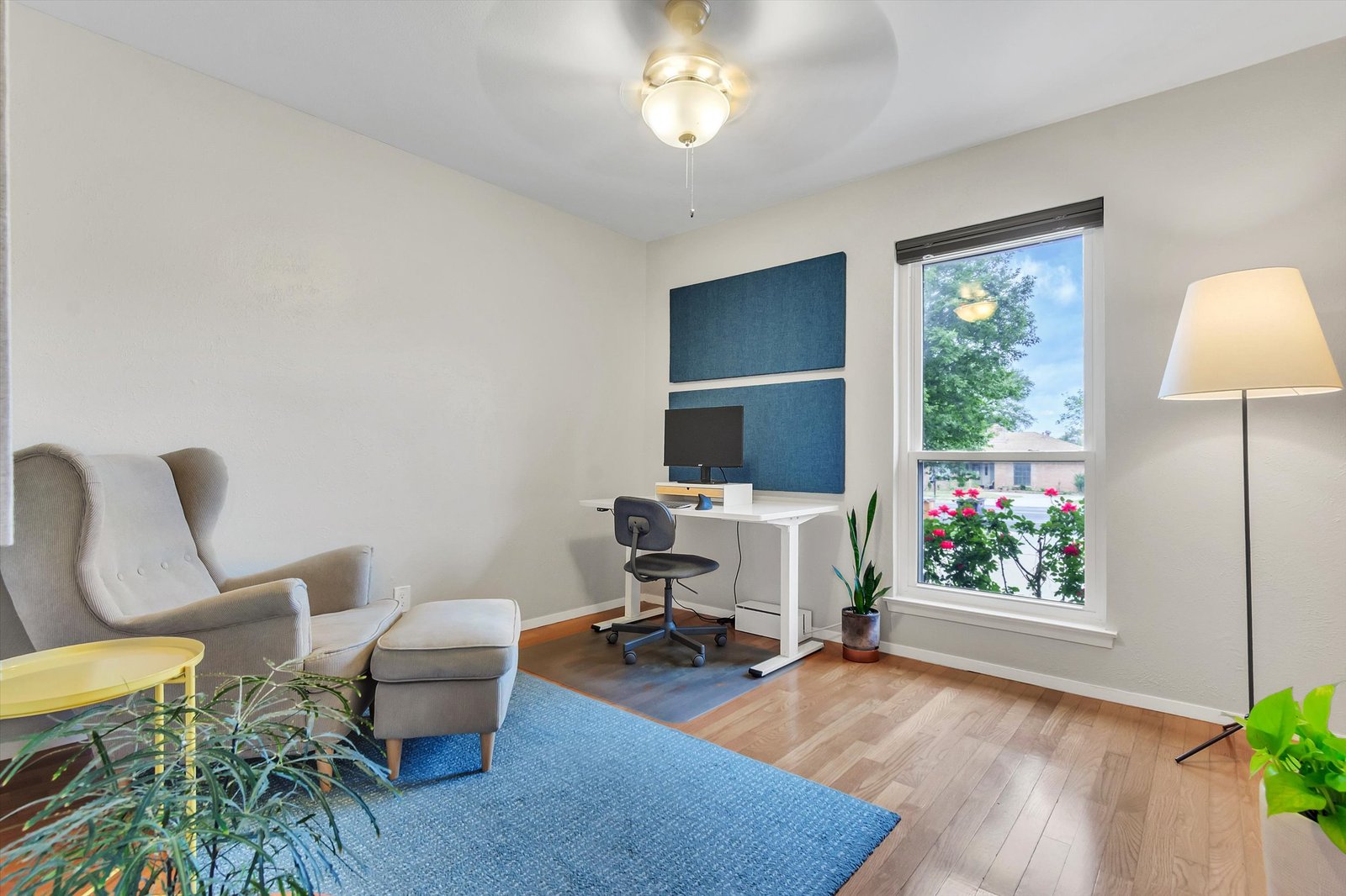 Bedroom 3 used as office — wingback chair, standing desk with monitor, blue rug, hardwood floors, and window overlooking front yard