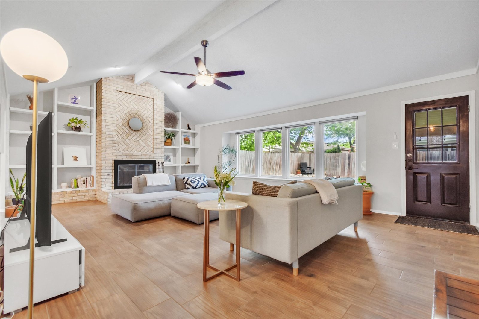 Living room from fireplace side showing mahogany front door, wall of windows, vaulted ceiling with beam, and open floor plan