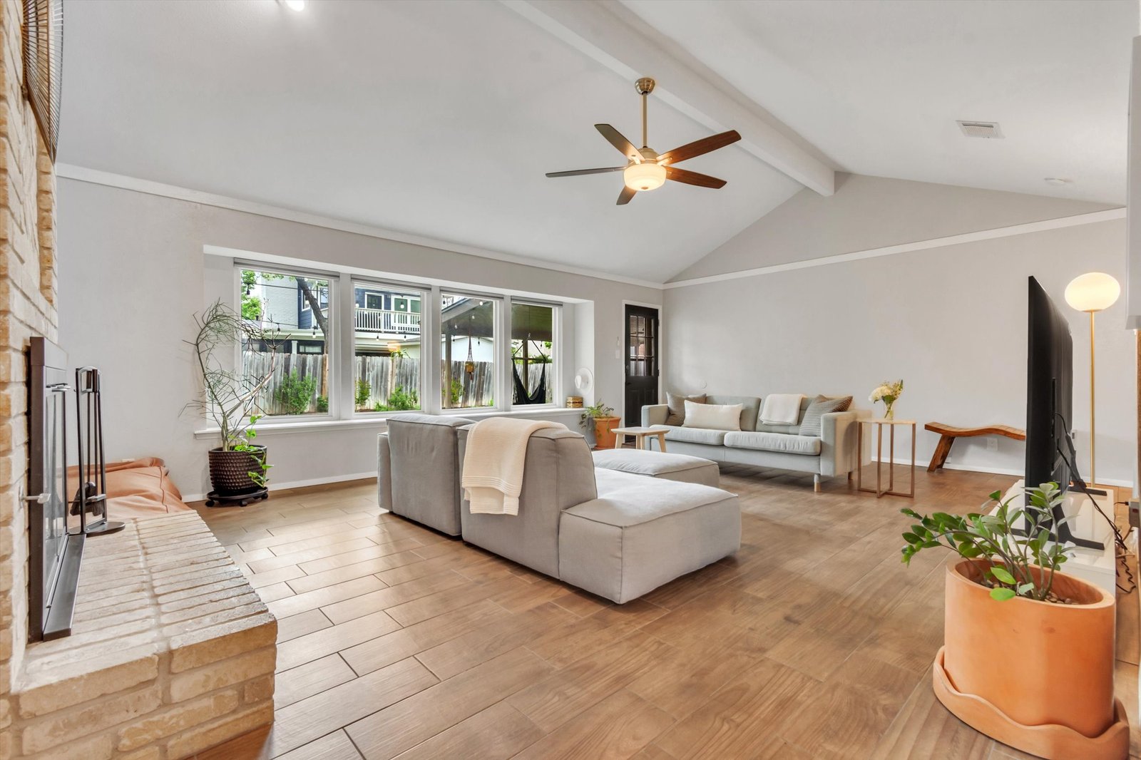 Living room wide angle showing wall of windows overlooking backyard, vaulted ceiling with beam, fireplace, and open layout to second living area