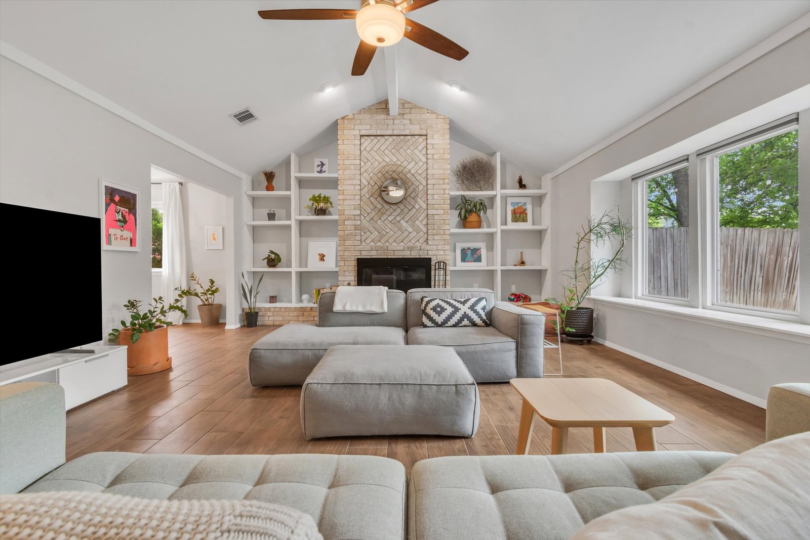 Living room with herringbone brick fireplace flanked by built-in bookcases, vaulted ceiling with beam, ceiling fan, and sectional sofa on tile floors