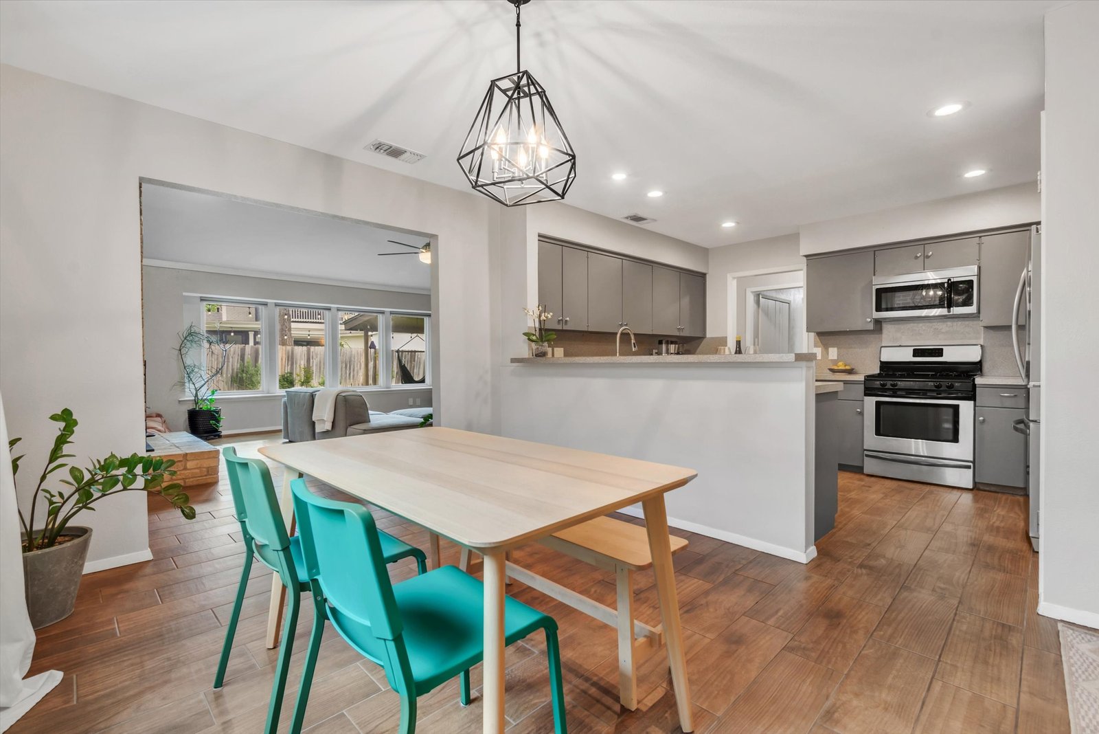 Dining area with geometric chandelier, teal chairs, and bench seat table — open to kitchen with breakfast bar and living room beyond