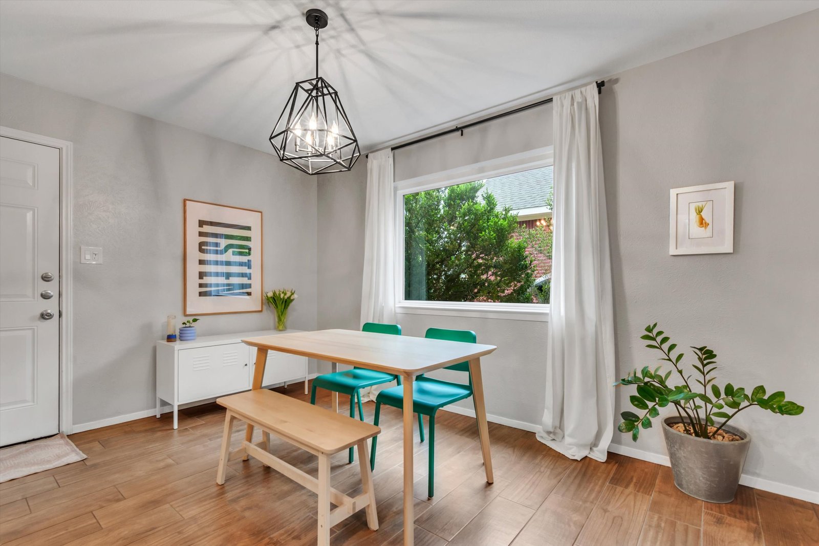 Dining room with large window, white curtains, geometric chandelier, teal chairs and wood bench at table, plants and art