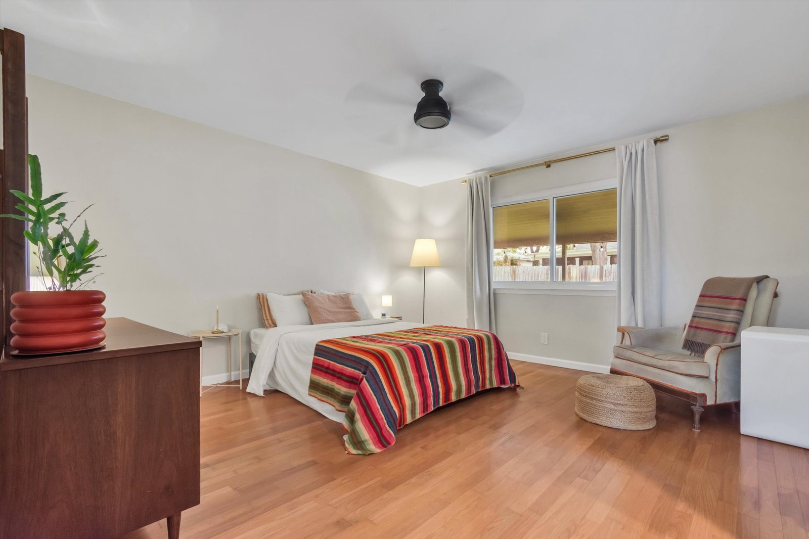 Spacious primary bedroom with hardwood floors, ceiling fan, mid-century dresser, and window looking out to backyard