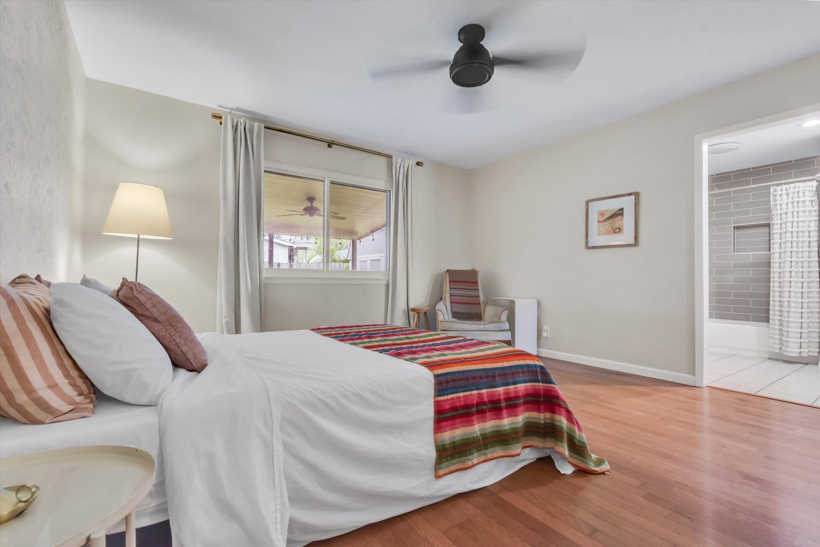 Primary bedroom from opposite angle showing bed, striped blanket, ceiling fan, window to covered patio, and ensuite bathroom entry with subway tile visible