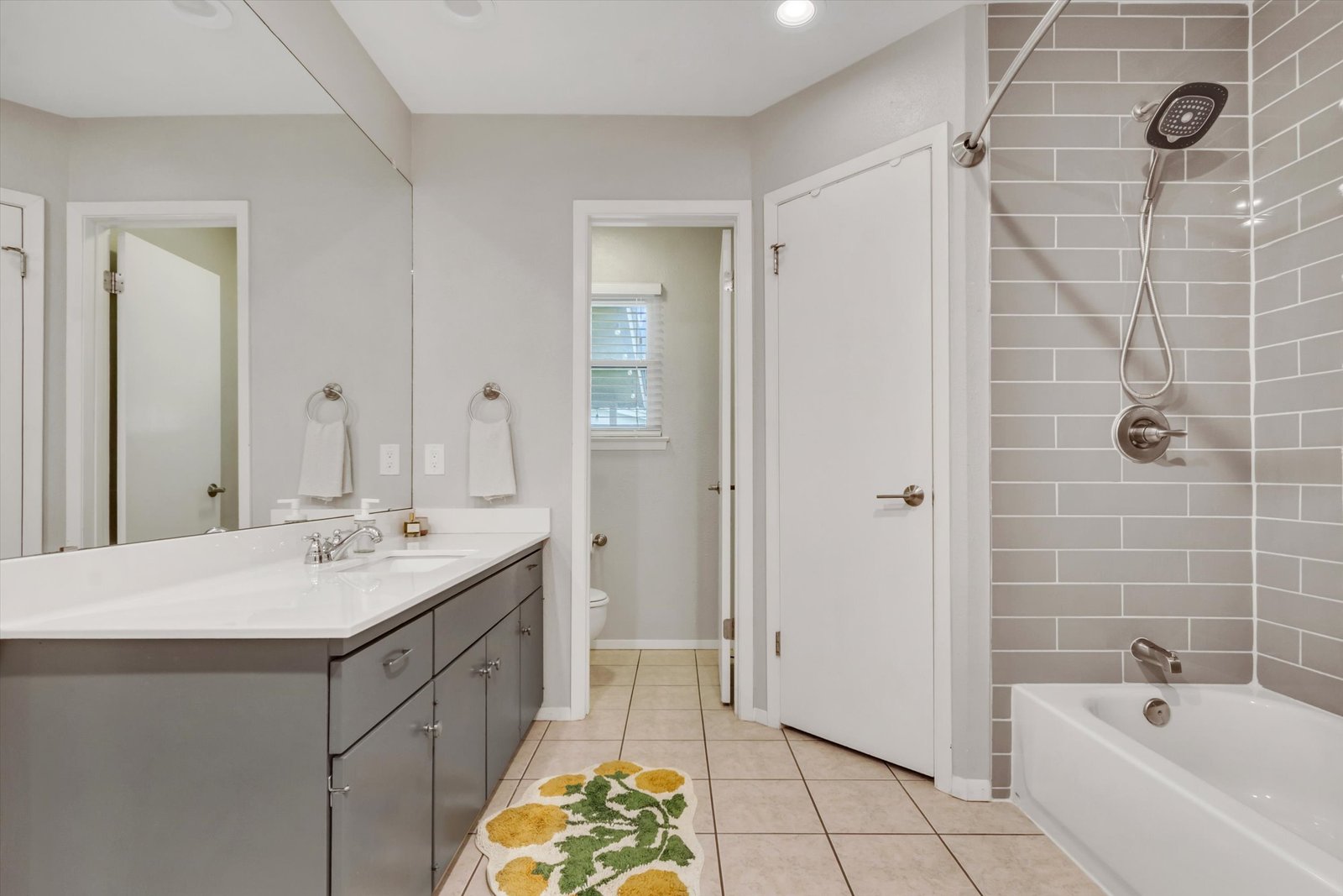 Primary bath vanity side — gray cabinets, white quartz countertop, large mirror, toilet room with window, subway tile shower visible