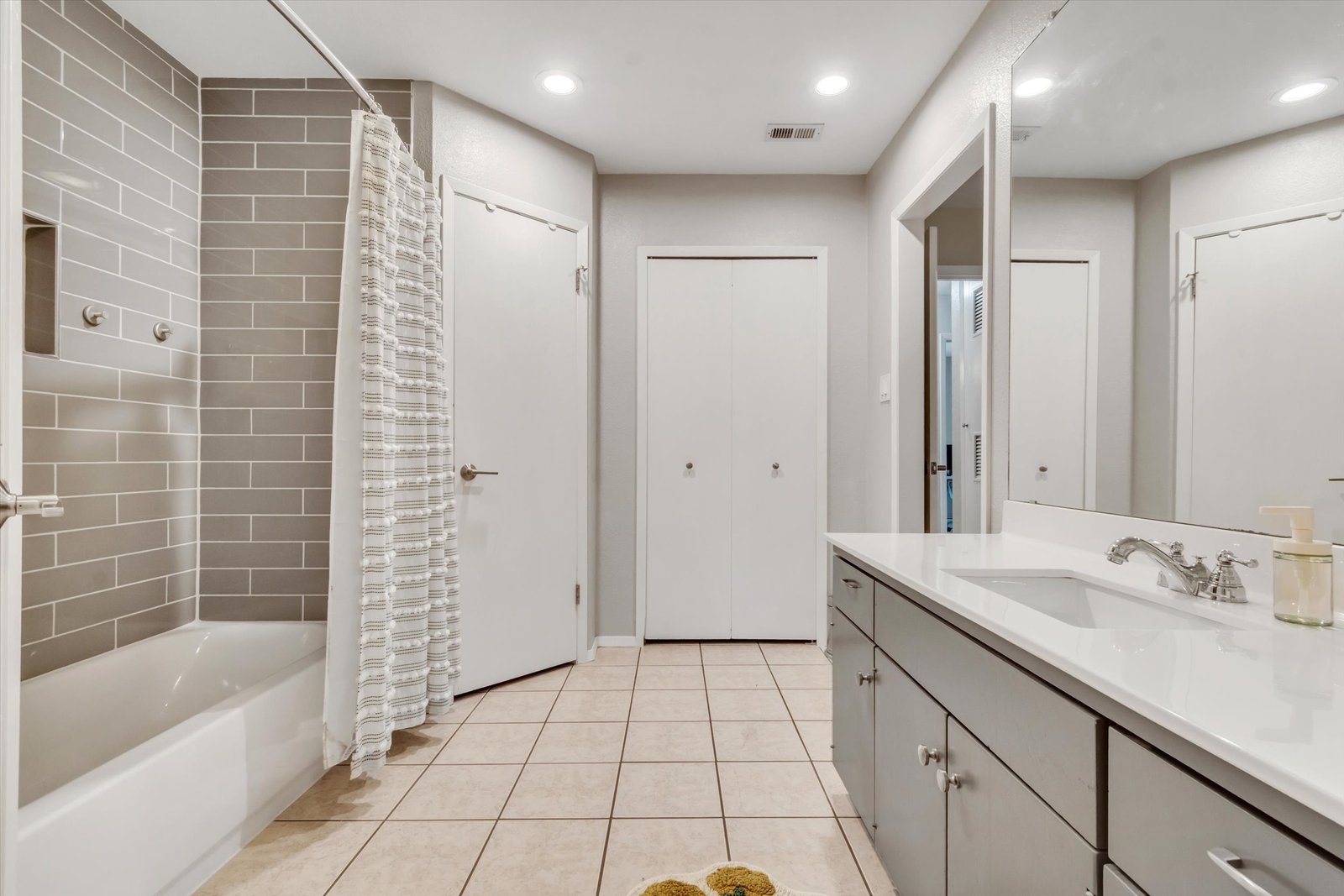 Primary ensuite bathroom — gray vanity with white countertop, subway tile tub/shower surround, bi-fold closet doors, recessed lighting