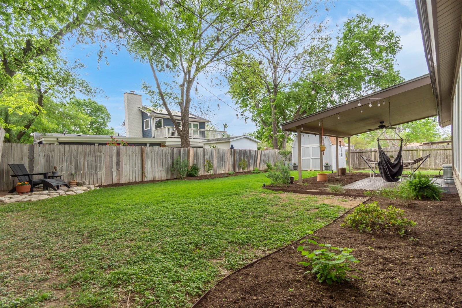 Backyard from garden beds — view of covered patio, string lights, mature trees, green lawn, and plantings
