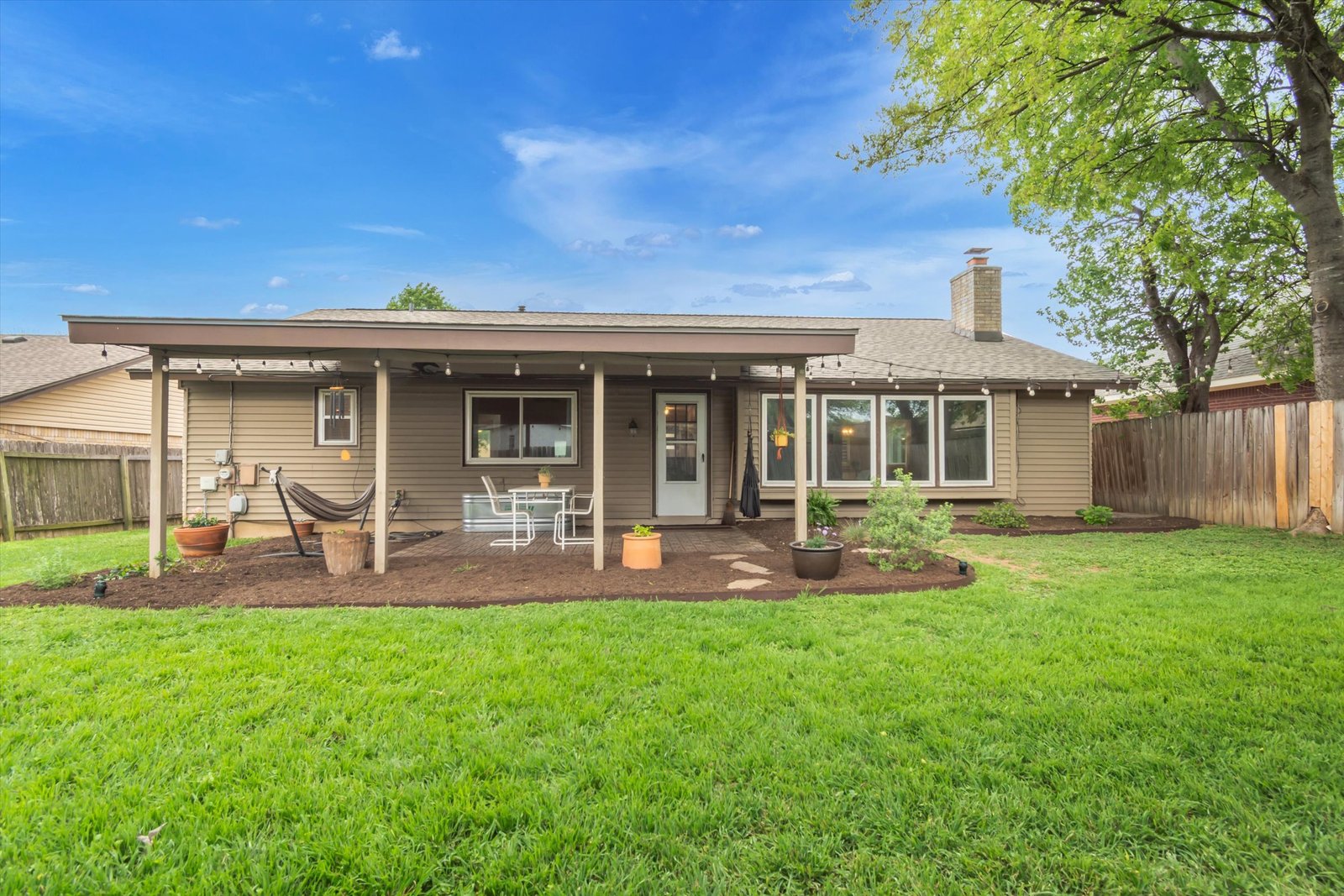 Rear exterior of home — covered patio with string lights, hammock, dining table, large windows, chimney, and green backyard