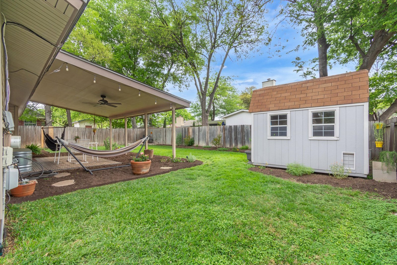 Backyard showing covered patio with hammock, ceiling fan, storage shed with shingled roof, mature trees, and fenced yard