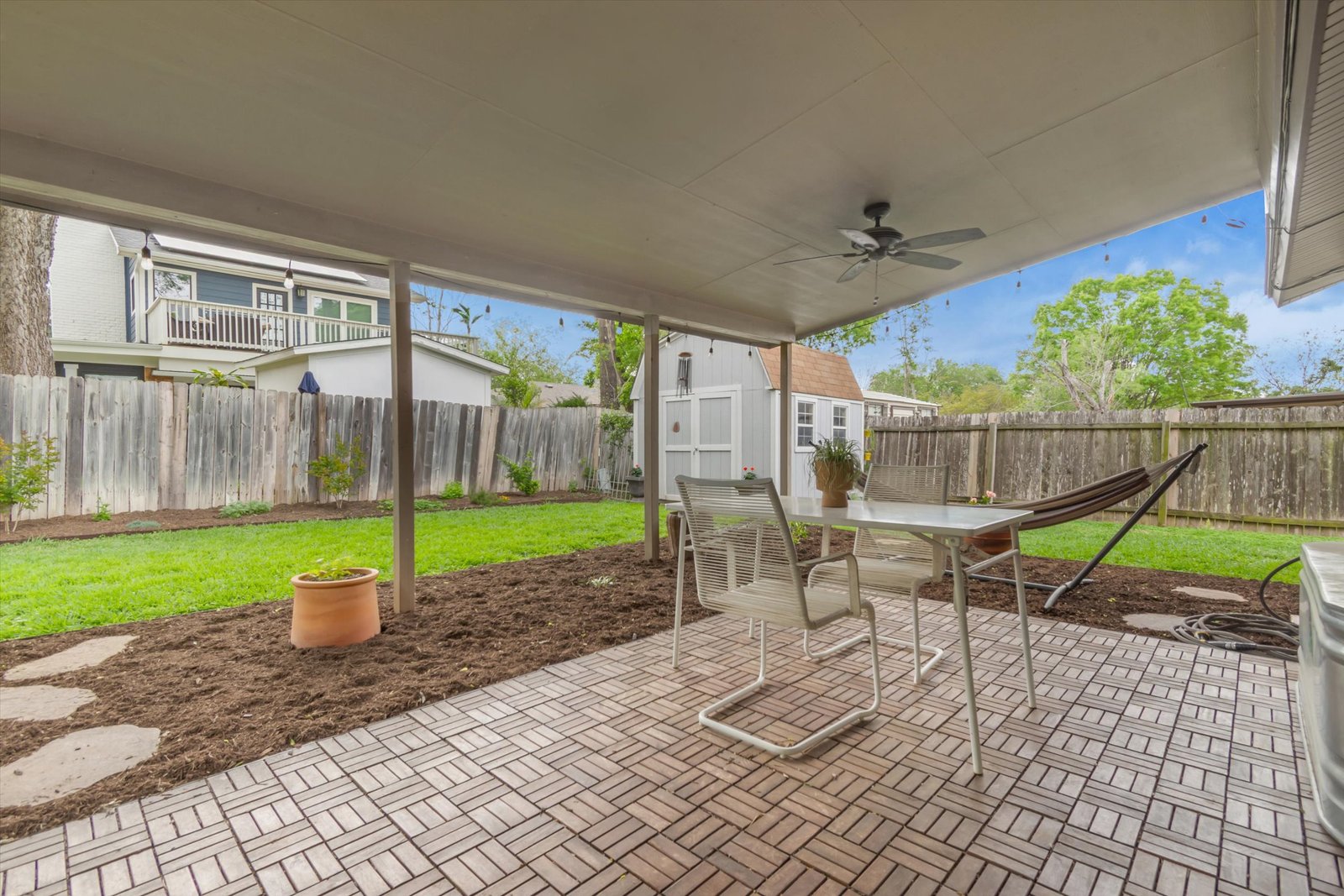 Under the covered patio — ceiling fan, dining table with chairs, brick pavers, looking out to fenced backyard with shed and hammock