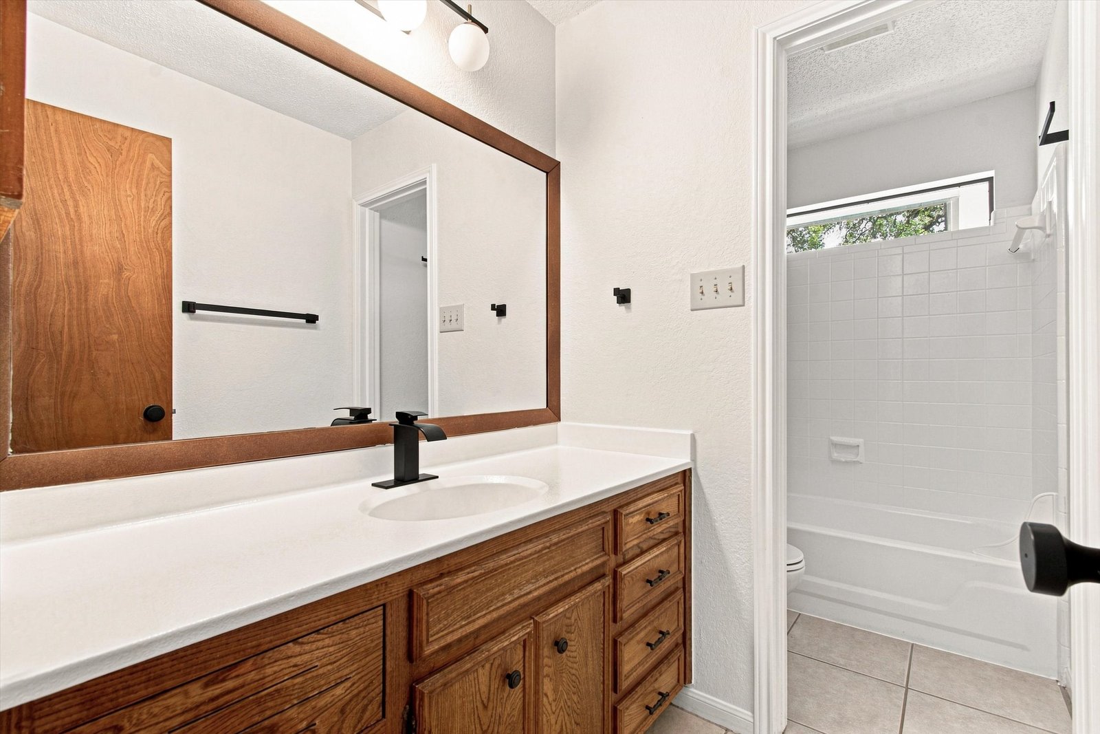 Guest bathroom with wood vanity, framed mirror, matte black fixtures, and tub-shower